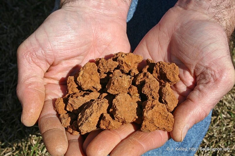 Two hands holding chunky red-brown clay soil aggregates